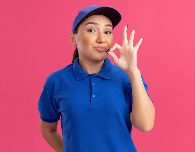 Young delivery woman in blue uniform and cap looking at front making silence gesture with fingers like closing mouth with a zipper standing over pink wall