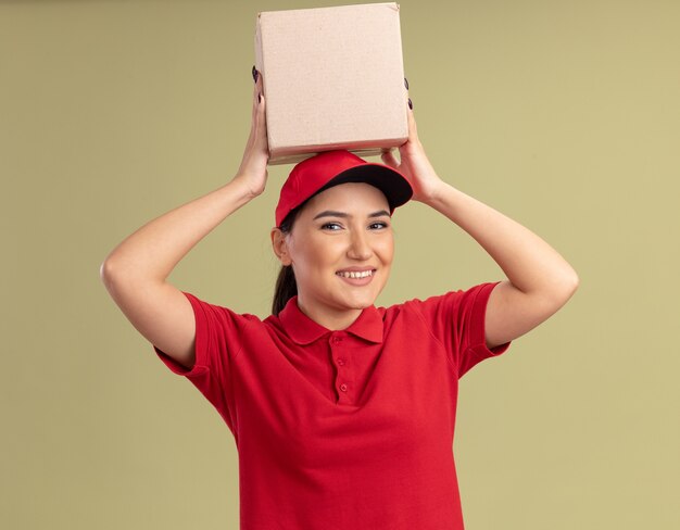 Young delivery woman in red uniform and cap holding cardboard box over her head looking at front smiling cheerfully standing over green wall