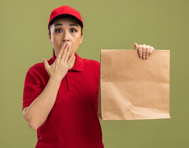 Young delivery woman in red uniform and cap holding paper package   being shocked covering mouth with hand  