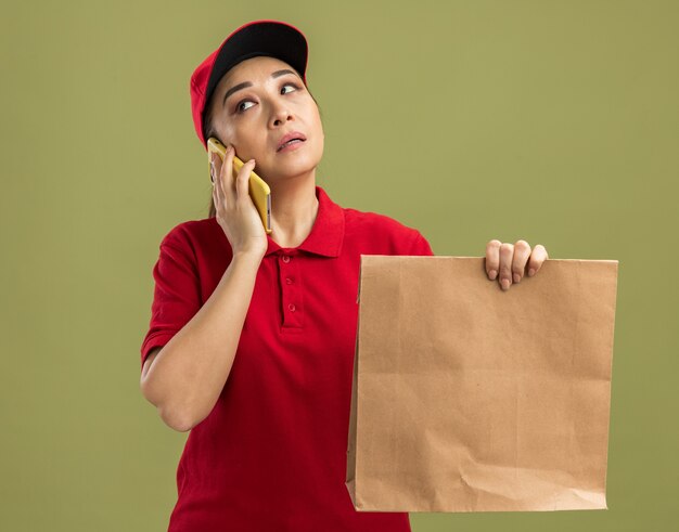 Young delivery woman in red uniform and cap holding paper package looking confused while talking on mobile phone standing over green wall