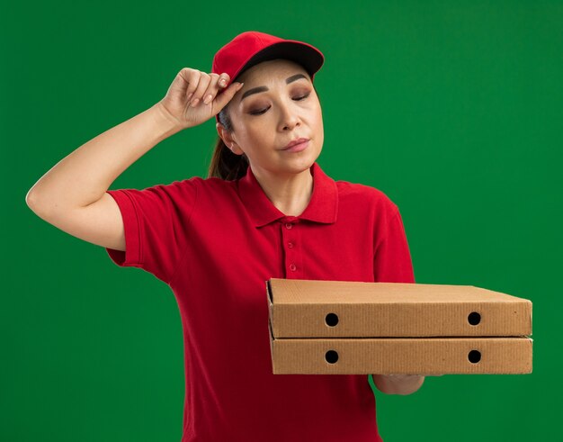 Young delivery woman in red uniform and cap holding pizza boxes looking down with sad expression standing over green wall