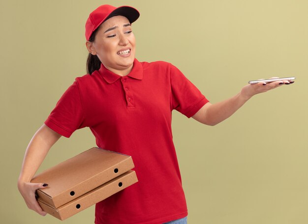 Young delivery woman in red uniform and cap holding pizza boxes looking at her smartphone in hand with confuse expression standing over green wall