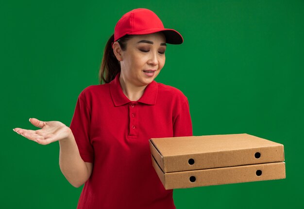 Young delivery woman in red uniform and cap holding pizza boxeslooking confused with arm out standing over green wall