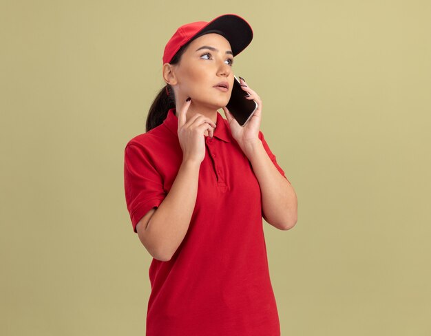 Young delivery woman in red uniform and cap talking on mobile phone with serious face standing over green wall