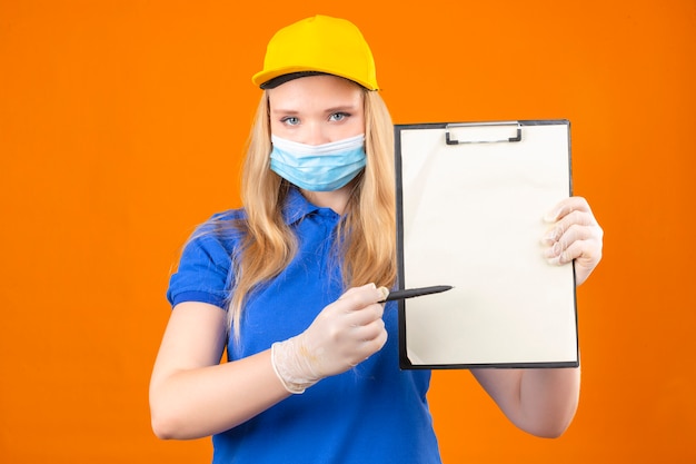 Free photo young delivery woman wearing blue polo shirt and yellow cap in medical protective mask standing with clipboard pointing with pen asking for signature with serious face over isolated dark yellow