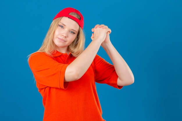 Young delivery woman wearing orange polo shirt and red cap gesturing with clasped looking confident and proud over isolated blue background