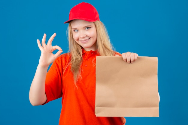 Young delivery woman wearing orange polo shirt and red cap holding paper package doing ok sign smiling friendly over isolated blue background