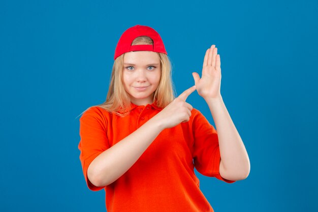 Young delivery woman wearing orange polo shirt and red cap pointing to open pal hand looking at camera with smile on face over isolated blue background