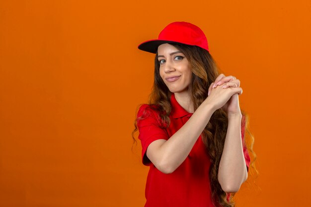 Young delivery woman wearing red polo shirt and cap gesturing with clasped looking confident and proud over isolated orange background