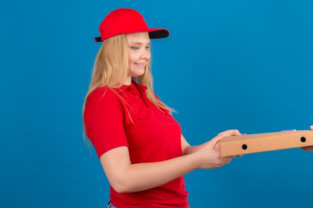 Young delivery woman wearing red polo shirt and cap giving pizza box to a customer smiling friendly over isolated blue background