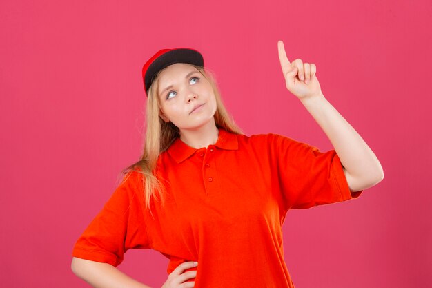 Young delivery woman wearing red polo shirt and cap looking up and pointing to something with index finger over isolated pink background