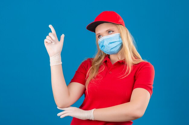 Young delivery woman wearing red polo shirt and cap in medical protective mask pointing with the index finger a great idea over isolated blue background