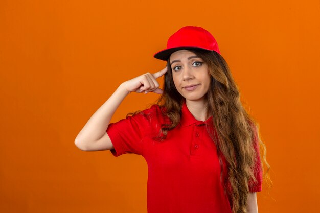 Young delivery woman wearing red polo shirt and cap pointing temple with finger skeptic look over isolated orange background