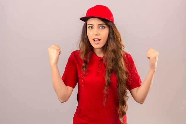 Free photo young delivery woman wearing red polo shirt and cap raising fists after a victory happy face winner concept over isolated white background