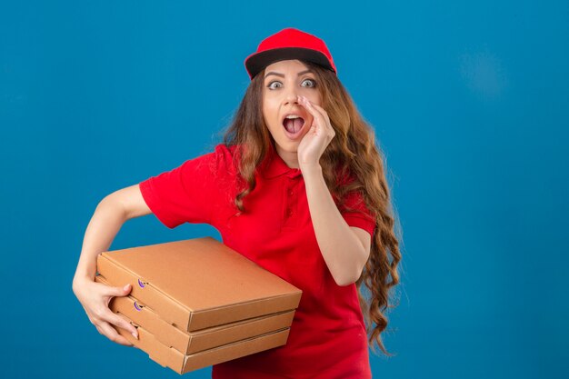 Young delivery woman wearing red polo shirt and cap standing with pizza boxes shouting to side with hand on mouth looking surprised over isolated blue background