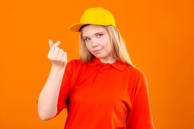 Young delivery woman wearing red polo shirt and yellow cap doing money gesture smiling over isolated orange background