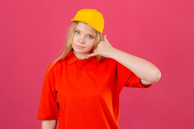 Young delivery woman wearing red polo shirt and yellow cap making call me gesture looking confident over isolated pink background