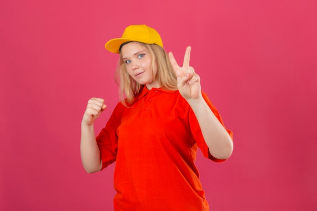 Free photo young delivery woman wearing red polo shirt and yellow cap smiling raising fist and showing victory sign winner concept over isolated pink background