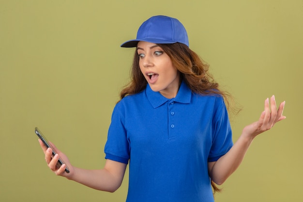 Free photo young delivery woman with curly hair wearing blue polo shirt and cap looking at screen of smartphone surprised with hand raised over isolated green background