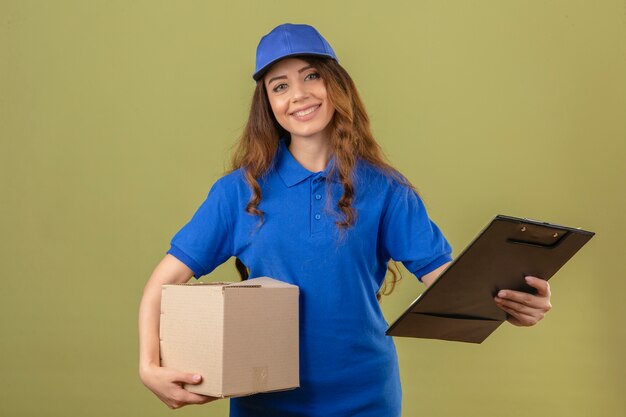 Young delivery woman with curly hair wearing blue polo shirt and cap standing with cardboard box and clipboard smiling friendly over isolated green background