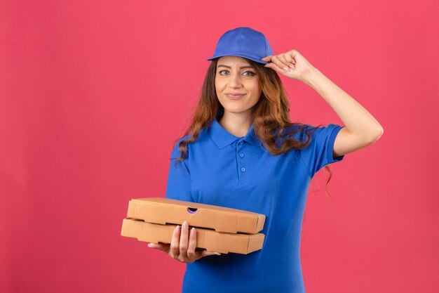 Young delivery woman with curly hair wearing blue polo shirt and cap standing with pizza boxes saluting looking confident over isolated pink background