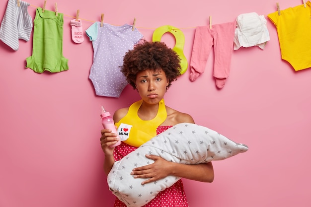 Young displeased tired mother holds baby wrapped in blanket holds feeding bottle wears bib around neck going to feed newborn has much work about house. Mom nursing small daughter. One parent family