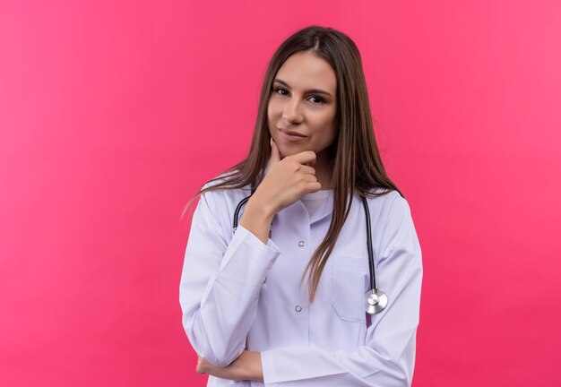  young doctor girl wearing stethoscope medical gown put her hand on chin on isolated pink wall