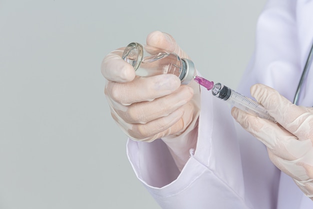 Young doctor is  holding  hypodermic syringe with  vaccine vial  rubber gloves on gray wall.