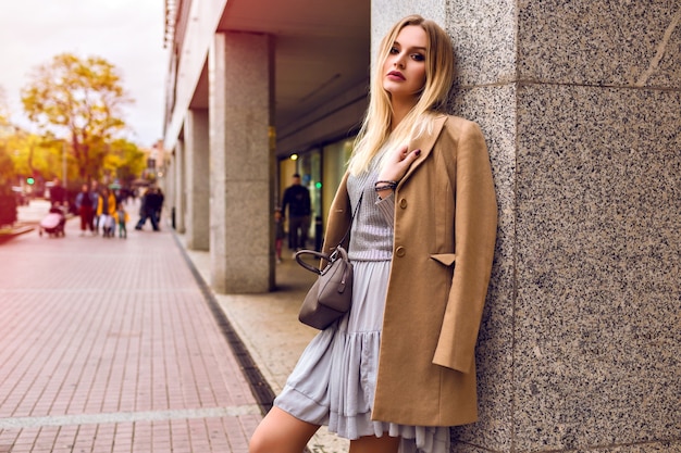 Young elegant woman posing on the street near shopping center, glamour trendy outfits, beige coat , silver sweater and dress, spring time, natural beauty