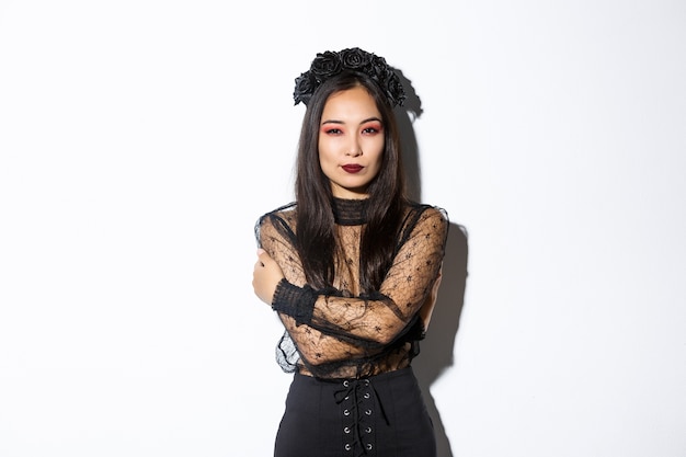 Free photo young elegant woman in stylish gothic dress and wreath, embracing her body and looking sassy at camera, wearing halloween costume of undead widow, standing over white background.