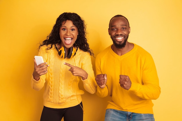 Young emotional african-american man and woman in bright casual clothes on yellow background. Beautiful couple. Concept of human emotions, facial expession, relations. Shocked pointing on smartphone.