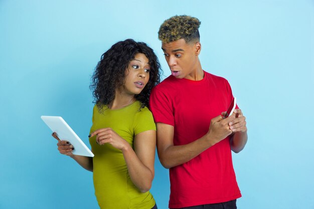 Young emotional african-american man and woman in colorful clothes using gadgets, peeping on screens on blue b