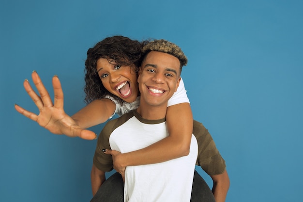 Young emotional african-american man and woman in white casual clothes posing on blue background. Beautiful couple. Concept of human emotions, facial expession, relations, ad. Hugging, laughting.