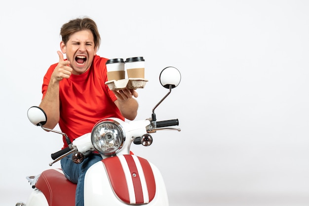 Free photo young emotional ambitious courier guy in red uniform sitting on scooter holding orders and making ok gesture on yellow wall