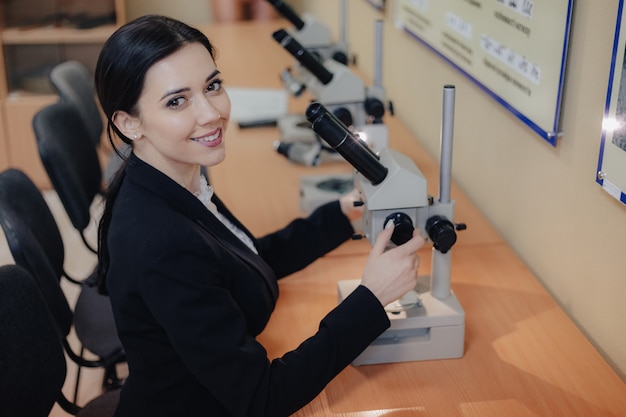 Young emotional attractive girl sitting at the table and working with a microscope in a modern office or audience