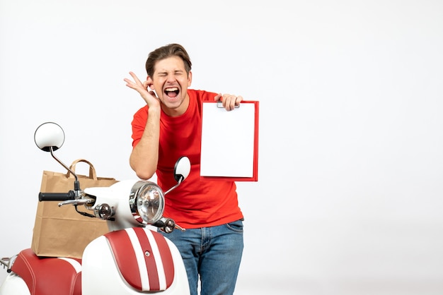 Free Photo young emotional nervous delivery guy in red uniform standing near scooter showing document closing one of his ear on white wall