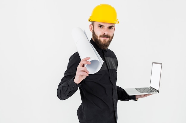 A young engineer holding rolledup blueprint and open laptop against white backdrop