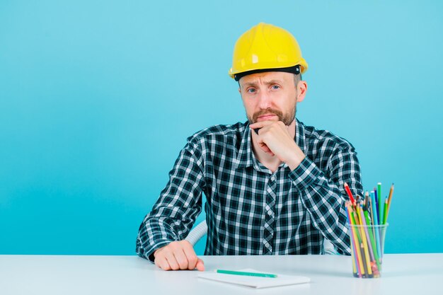 Young engineer man is looking at camera by holding hand on chin on blue background