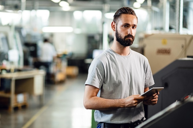 Young engineer using touchpad while operating a CNC machine in a factory and looking at camera