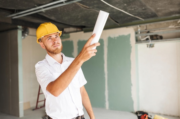 Free photo young engineer in white shirt and yellow hardhat holding plan of new apartment in hand thoughtfully looking on ceiling at work