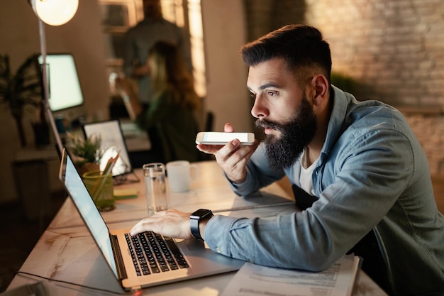 Young entrepreneur reading an email on computer while recording voice message on smart phone during late night work in the office