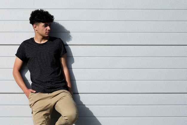 Young ethnic curly man with hands in pockets leaning on grey wall