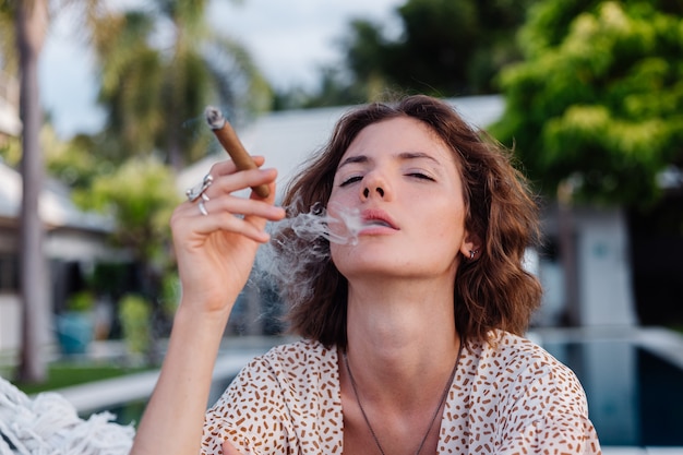 Young european woman smoking cigar lying on hammock outside tropical luxury villa hotel, sunset natural light