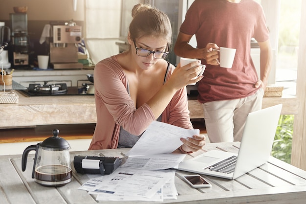 Free photo young family dealing financial issues. serious wife in spectacles sitting in front of laptop computer, looking through bills, holding cup