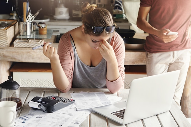 Young family facing debt problems. Stressed woman holding her head in frustration, doing bank accounts and making necessary calculations using laptop and calculator