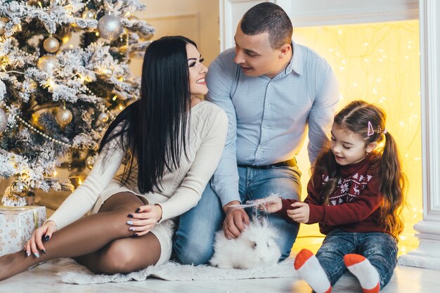 Young family poses before a shiny Christmas tree in a cosy luxury room