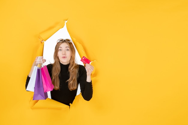 Free photo young fashion woman holding shopping bags and credit card through torn paper hole in the wall