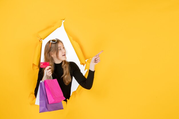 Young fashion woman holding shopping bags and credit card through torn paper hole in the wall