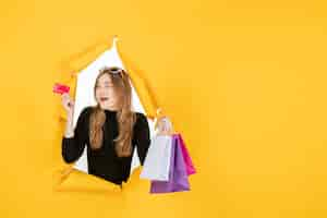 Free photo young fashion woman holding shopping bags and credit card through torn paper hole in the wall