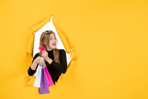 Free photo young fashion woman holding shopping bags and credit card through torn paper hole in the wall